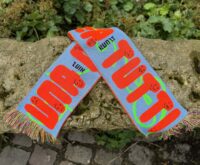 Ein blauer Fanschal mit orangefarbenen Schriftzügen "UNA CASA PER TUTTI" und stilisierten Gesichtern ist auf einem felsigen Untergrund abgebildet. In schwarz steht Hinz und Kunzt zwischen den Worten