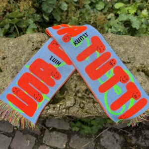 Ein blauer Fanschal mit orangefarbenen Schriftzügen "UNA CASA PER TUTTI" und stilisierten Gesichtern ist auf einem felsigen Untergrund abgebildet. In schwarz steht Hinz und Kunzt zwischen den Worten