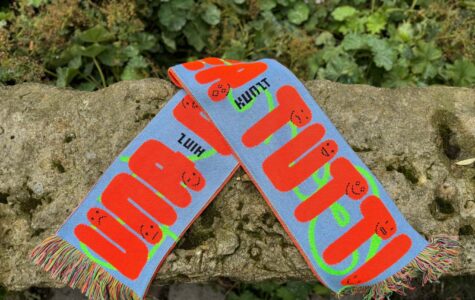 Ein blauer Fanschal mit orangefarbenen Schriftzügen "UNA CASA PER TUTTI" und stilisierten Gesichtern ist auf einem felsigen Untergrund abgebildet. In schwarz steht Hinz und Kunzt zwischen den Worten