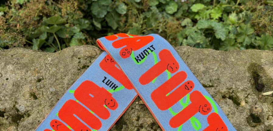 Ein blauer Fanschal mit orangefarbenen Schriftzügen "UNA CASA PER TUTTI" und stilisierten Gesichtern ist auf einem felsigen Untergrund abgebildet. In schwarz steht Hinz und Kunzt zwischen den Worten