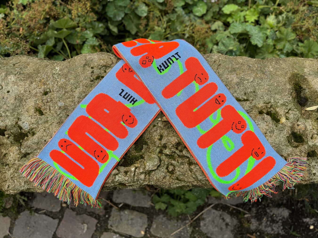 Ein blauer Fanschal mit orangefarbenen Schriftzügen "UNA CASA PER TUTTI" und stilisierten Gesichtern ist auf einem felsigen Untergrund abgebildet. In schwarz steht Hinz und Kunzt zwischen den Worten