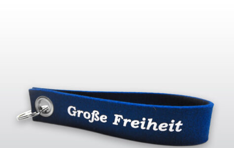 Das abgebildete Objekt ist ein dunkelblauer Schlüsselanhänger mit weißer Aufschrift „Große Freiheit“. Auf der anderen Seite befindet sich die Aufschrift „Hinz&Kunzt“ sowie der Herstellername „dekooop“.
