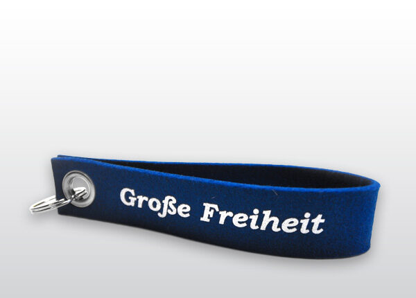 Das abgebildete Objekt ist ein dunkelblauer Schlüsselanhänger mit weißer Aufschrift „Große Freiheit“. Auf der anderen Seite befindet sich die Aufschrift „Hinz&Kunzt“ sowie der Herstellername „dekooop“.