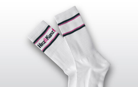 Das Bild zeigt ein Paar weiße Socken. Sie haben schwarze und magenta Streifen am Bund und tragen den Schriftzug "Hinz&Kunzt".