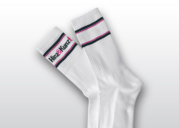 Das Bild zeigt ein Paar weiße Socken. Sie haben schwarze und magenta Streifen am Bund und tragen den Schriftzug "Hinz&Kunzt".