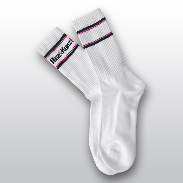 Das Bild zeigt ein Paar weiße Socken. Sie haben schwarze und magenta Streifen am Bund und tragen den Schriftzug "Hinz&Kunzt".