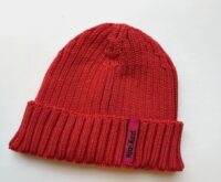 Die Abbildung zeigt eine Strickmütze in der Farbe Cranberry. Sie hat ein magentafarbenes Etikett, auf dem in schwarzer Schrift "Hinz&Kunzt" steht.