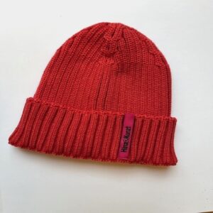 Die Abbildung zeigt eine Strickmütze in der Farbe Cranberry. Sie hat ein magentafarbenes Etikett, auf dem in schwarzer Schrift "Hinz&Kunzt" steht.