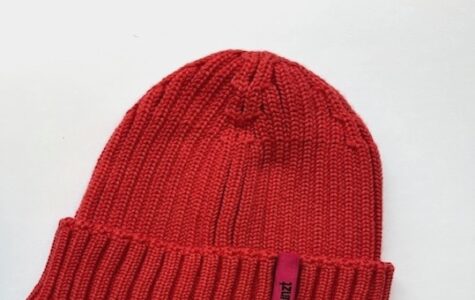 Die Abbildung zeigt eine Strickmütze in der Farbe Cranberry. Sie hat ein magentafarbenes Etikett, auf dem in schwarzer Schrift "Hinz&Kunzt" steht.