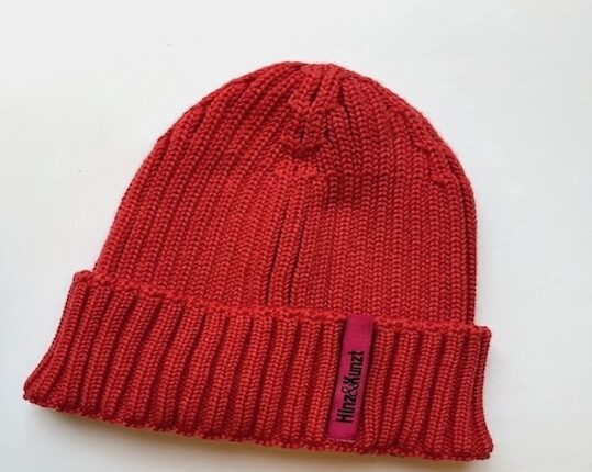 Die Abbildung zeigt eine Strickmütze in der Farbe Cranberry. Sie hat ein magentafarbenes Etikett, auf dem in schwarzer Schrift "Hinz&Kunzt" steht.