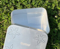 Das Bild zeigt eine offene, rechteckige Proviantdose aus Aluminium, die auf grünem Gras liegt. Die Dose ist silberfarben und weist auf dem Deckel ein Muster aus sternförmigen Perforationen auf. In der Mitte ist weiß Hinz&Kunzt eingestanzt.