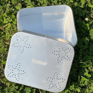 Das Bild zeigt eine offene, rechteckige Proviantdose aus Aluminium, die auf grünem Gras liegt. Die Dose ist silberfarben und weist auf dem Deckel ein Muster aus sternförmigen Perforationen auf. In der Mitte ist weiß Hinz&Kunzt eingestanzt.