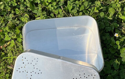 Das Bild zeigt eine offene, rechteckige Proviantdose aus Aluminium, die auf grünem Gras liegt. Die Dose ist silberfarben und weist auf dem Deckel ein Muster aus sternförmigen Perforationen auf. In der Mitte ist weiß Hinz&Kunzt eingestanzt.