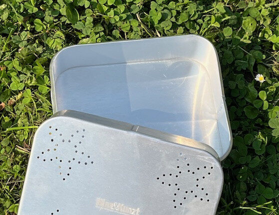 Das Bild zeigt eine offene, rechteckige Proviantdose aus Aluminium, die auf grünem Gras liegt. Die Dose ist silberfarben und weist auf dem Deckel ein Muster aus sternförmigen Perforationen auf. In der Mitte ist weiß Hinz&Kunzt eingestanzt.