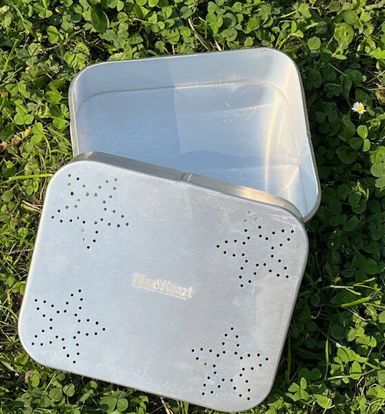 Das Bild zeigt eine offene, rechteckige Proviantdose aus Aluminium, die auf grünem Gras liegt. Die Dose ist silberfarben und weist auf dem Deckel ein Muster aus sternförmigen Perforationen auf. In der Mitte ist weiß Hinz&Kunzt eingestanzt.