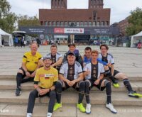 Das Deutsche Straßenfußball-Nationalteam sitzt auf einem Platz auf einer Treppe und grinst in die Kamera.