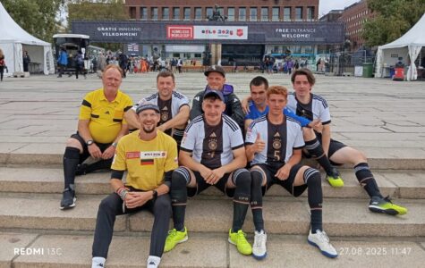 Das Deutsche Straßenfußball-Nationalteam sitzt auf einem Platz auf einer Treppe und grinst in die Kamera.