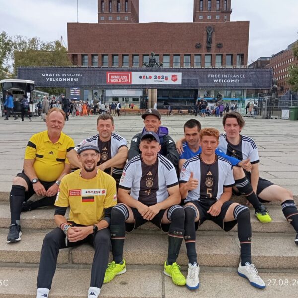 Das Deutsche Straßenfußball-Nationalteam sitzt auf einem Platz auf einer Treppe und grinst in die Kamera.