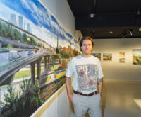 Ein Mann im weißen T-Shirt steht vor einem Gemälde und schaut in die Kamera