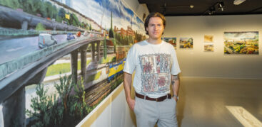Ein Mann im weißen T-Shirt steht vor einem Gemälde und schaut in die Kamera