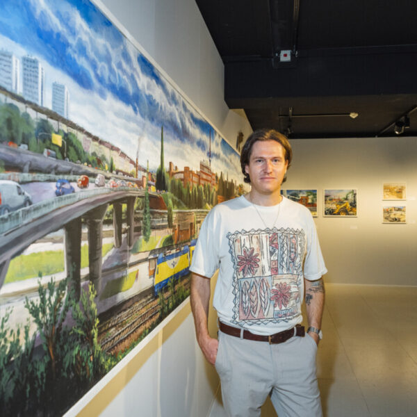Ein Mann im weißen T-Shirt steht vor einem Gemälde und schaut in die Kamera
