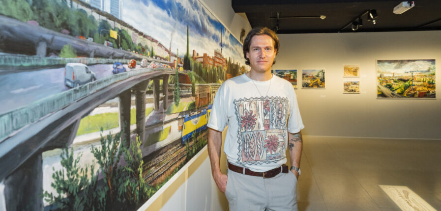 Ein Mann im weißen T-Shirt steht vor einem Gemälde und schaut in die Kamera