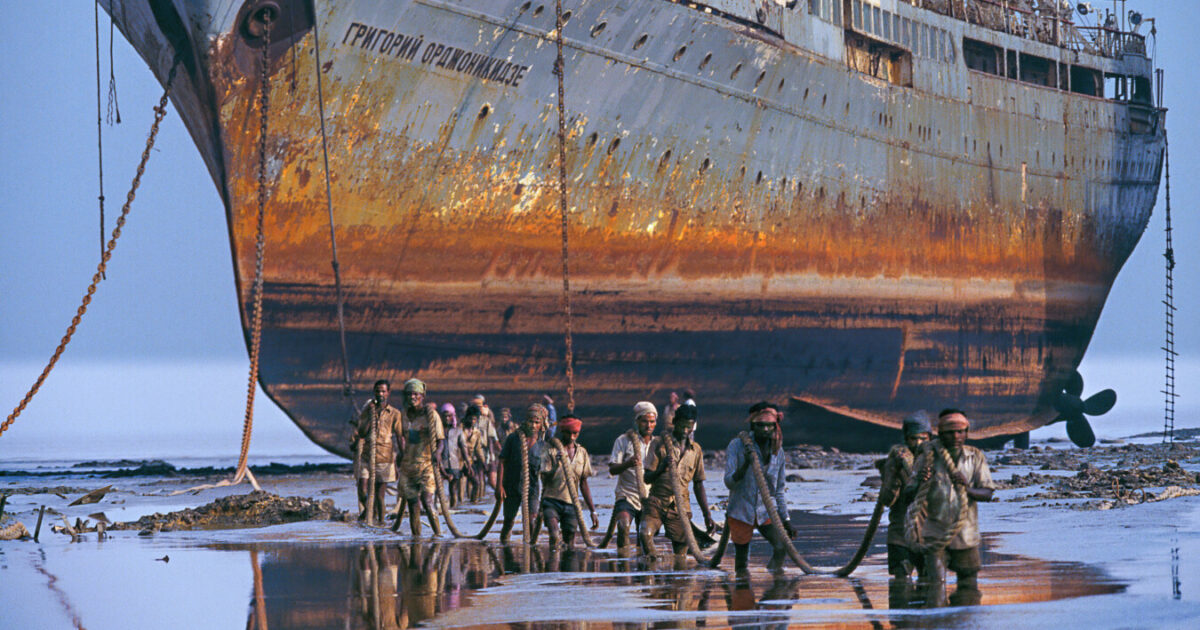Ein riesiges rostiges Schiff lieft trocken davor waten Männer durch knietiefes Wasser