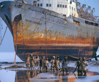 Ein riesiges rostiges Schiff lieft trocken davor waten Männer durch knietiefes Wasser