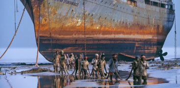 Ein riesiges rostiges Schiff lieft trocken davor waten Männer durch knietiefes Wasser