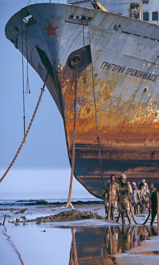 Ein riesiges rostiges Schiff lieft trocken davor waten Männer durch knietiefes Wasser