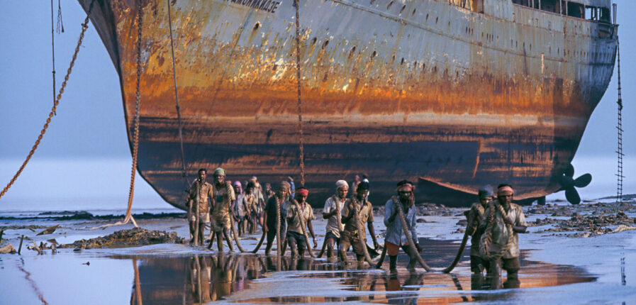 Ein riesiges rostiges Schiff lieft trocken davor waten Männer durch knietiefes Wasser