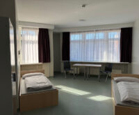 2 Betten und ein Schrank stehen in einem Raum. Im Hintergrund ist ein Tisch mit zwei Stühlen zu sehen.