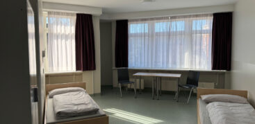 2 Betten und ein Schrank stehen in einem Raum. Im Hintergrund ist ein Tisch mit zwei Stühlen zu sehen.