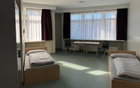 2 Betten und ein Schrank stehen in einem Raum. Im Hintergrund ist ein Tisch mit zwei Stühlen zu sehen.