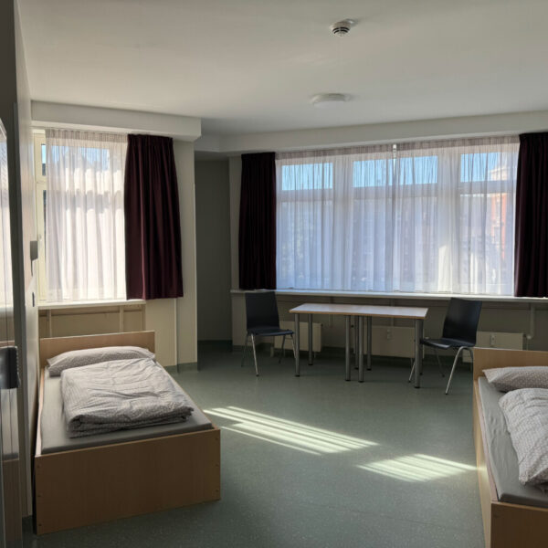 2 Betten und ein Schrank stehen in einem Raum. Im Hintergrund ist ein Tisch mit zwei Stühlen zu sehen.