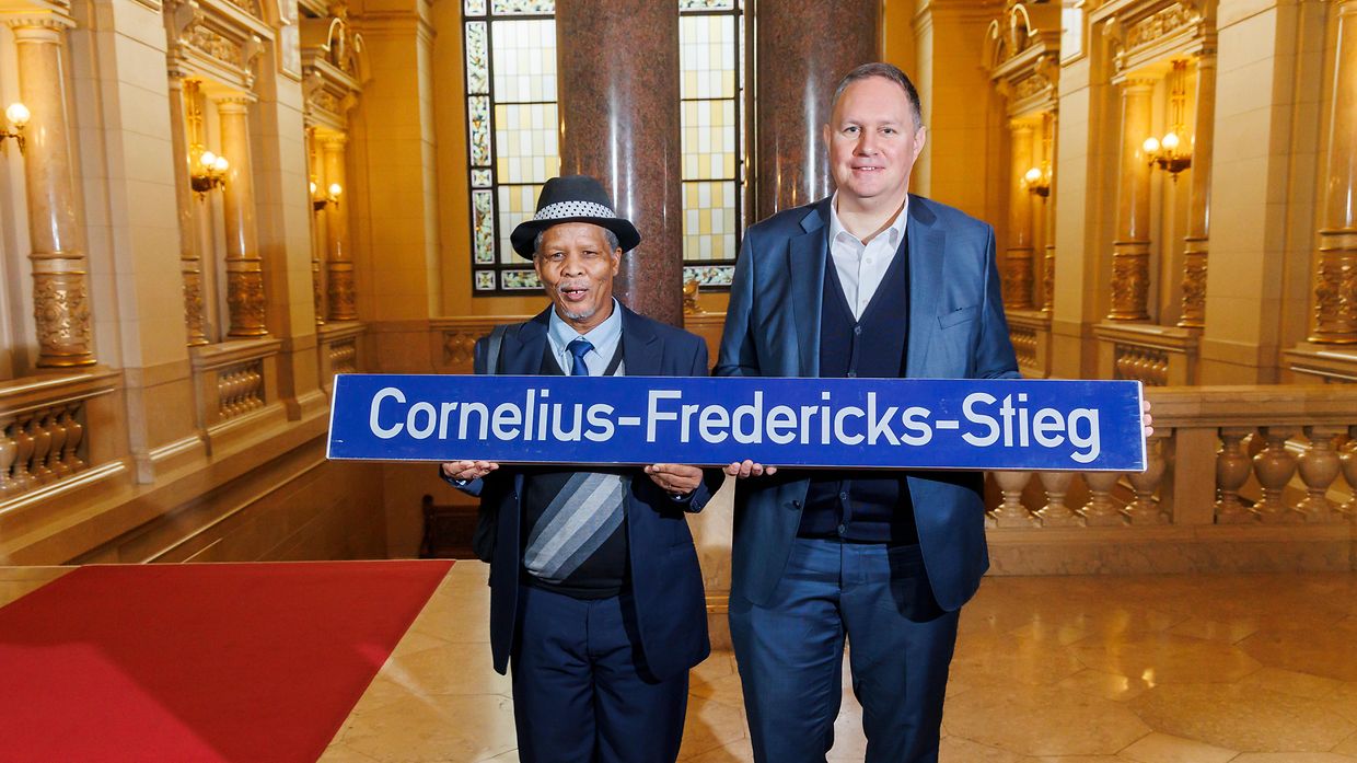 Zwei Männer stehen im Hamburger Rathaus und halten ein Straßenschild mit der Aufschrift "Cornelius-Fredericks-Stieg" hoch.