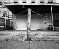 Schwarzweiß-Foto von einem Poller vor einem Ladengeschäft, dessen Rollo geschlossen ist