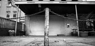 Schwarzweiß-Foto von einem Poller vor einem Ladengeschäft, dessen Rollo geschlossen ist