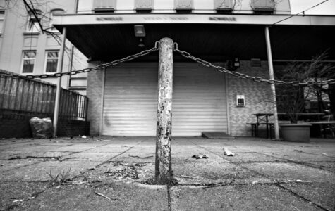 Schwarzweiß-Foto von einem Poller vor einem Ladengeschäft, dessen Rollo geschlossen ist