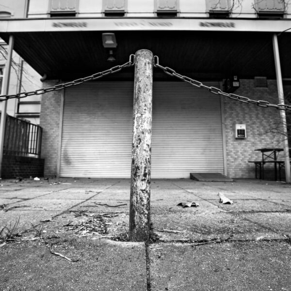 Schwarzweiß-Foto von einem Poller vor einem Ladengeschäft, dessen Rollo geschlossen ist