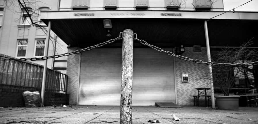 Schwarzweiß-Foto von einem Poller vor einem Ladengeschäft, dessen Rollo geschlossen ist