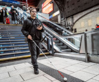 Ein Mann mit Taststock und Smartphone in der Hand folgt einem Leitstreifen im Bahnhofsgebäude