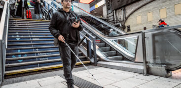 Ein Mann mit Taststock und Smartphone in der Hand folgt einem Leitstreifen im Bahnhofsgebäude
