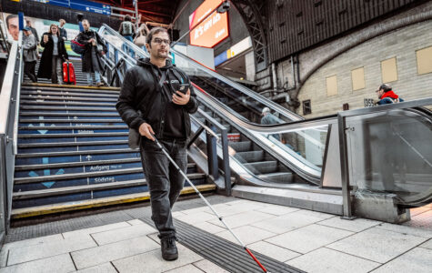 Ein Mann mit Taststock und Smartphone in der Hand folgt einem Leitstreifen im Bahnhofsgebäude