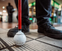 Füße und ein Taststock stehen auf einem Leitstreifen