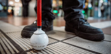 Füße und ein Taststock stehen auf einem Leitstreifen