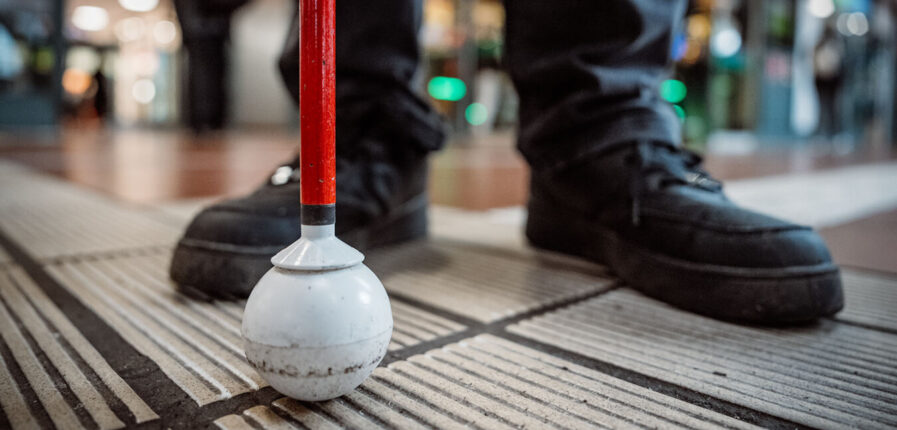 Füße und ein Taststock stehen auf einem Leitstreifen