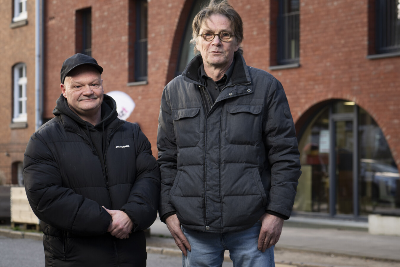 Ein Mann mit dunkler Kappe in schwarzer Jacke und ein Mann mit Brille und grauer Jahre stehen vor einem Rotklinkergebäude