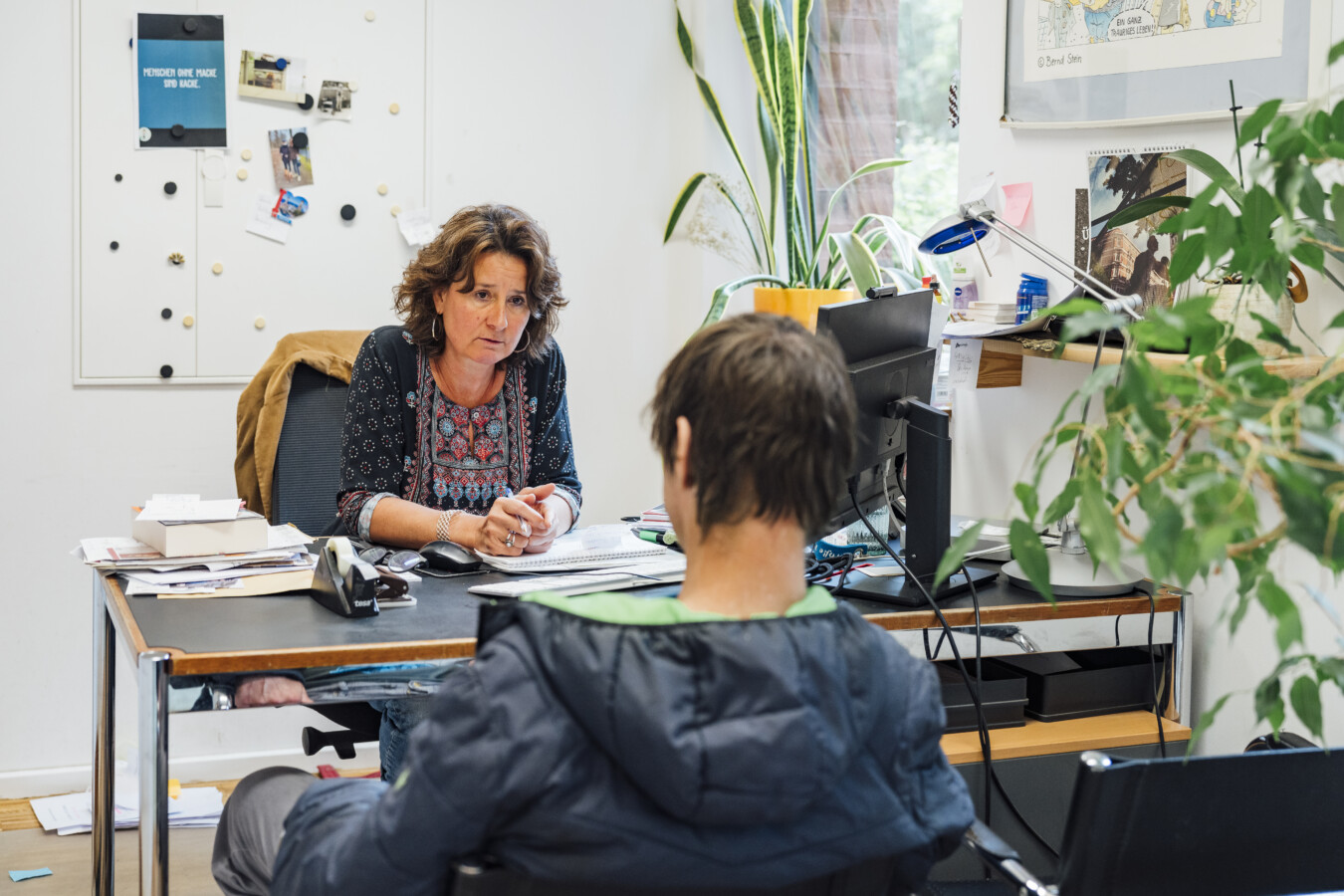 In einem Büro mit einigen Pflanzen ist eine dunkelhaarige Frau zu sehen. Sie spricht mit einem dunkelhaarigen Mann, der mit dem Rücken zur Kamera sitzt.
