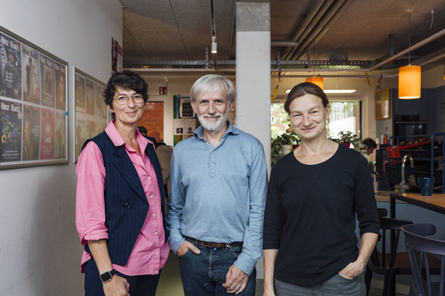 Zwei dunkelhaarige Frau und ein grauhaariger Mann (Mitte) stehen in einem Innenraum.
